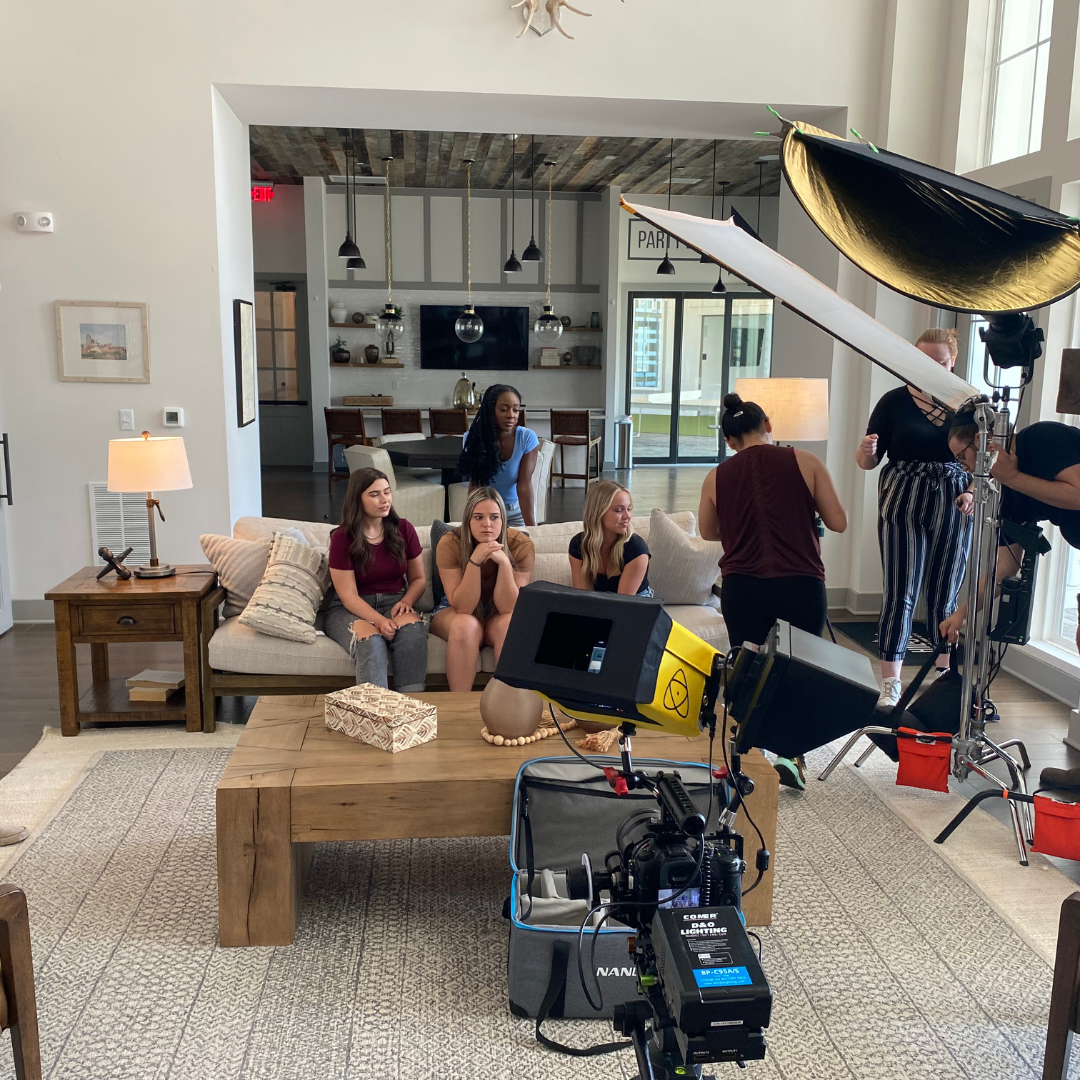 A film or photo shoot setup in a modern living room with four women sitting on a beige couch, while a crew member adjusts equipment. There are professional lighting and camera equipment, with a large softbox light on the right and a camera on a tripo