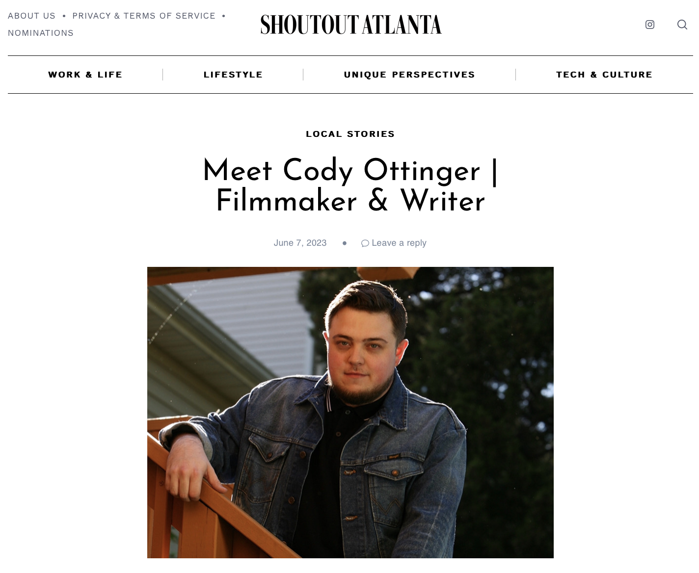 Young man with short dark hair and beard wearing a dark denim jacket, standing outdoors on a staircase with a house and trees in the background.