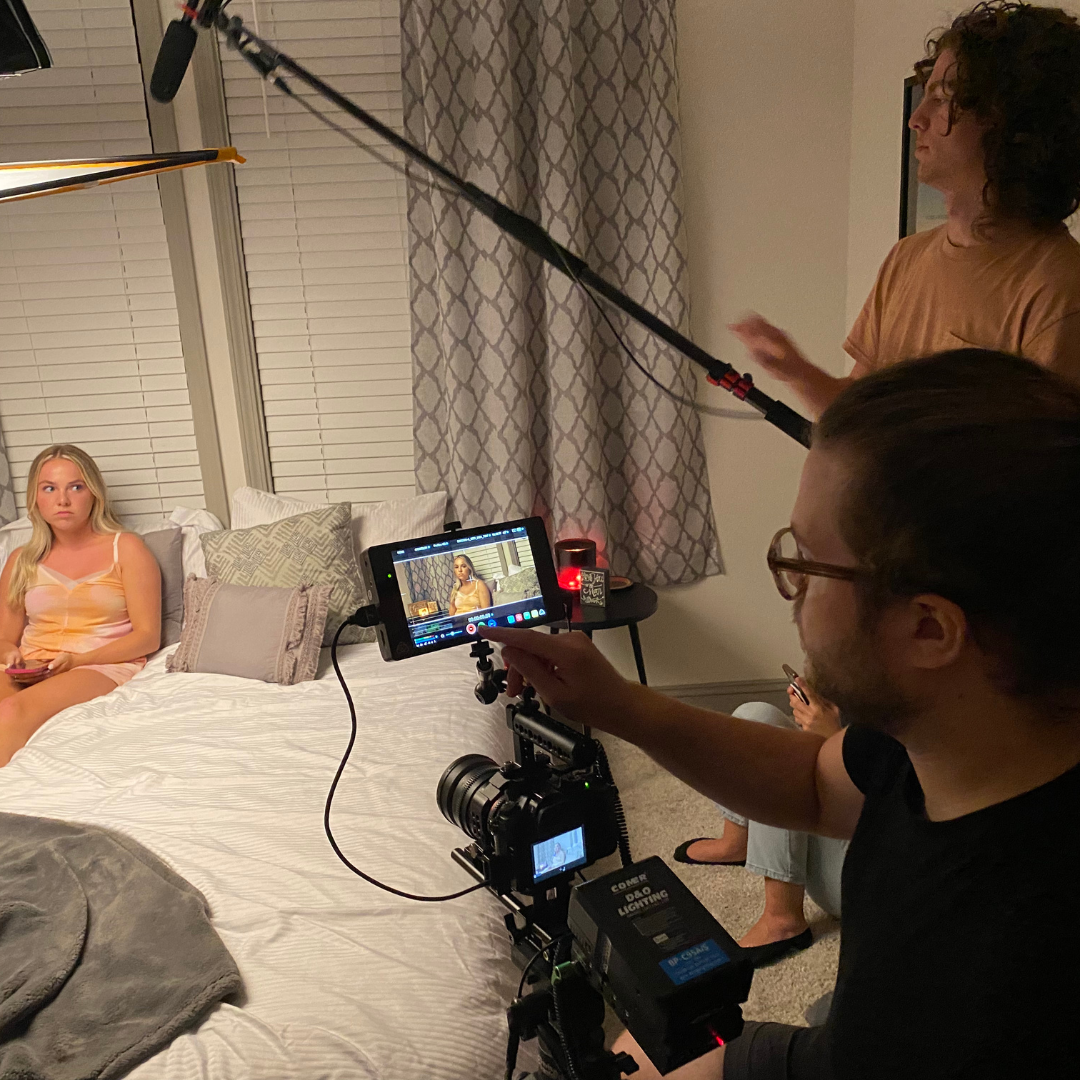 A woman sits on a bed with a serious expression while being filmed during a video shoot or interview. A crew member operates a camera and monitors the shot on a screen, a man holds a boom microphone above, and another person is partially visible in t