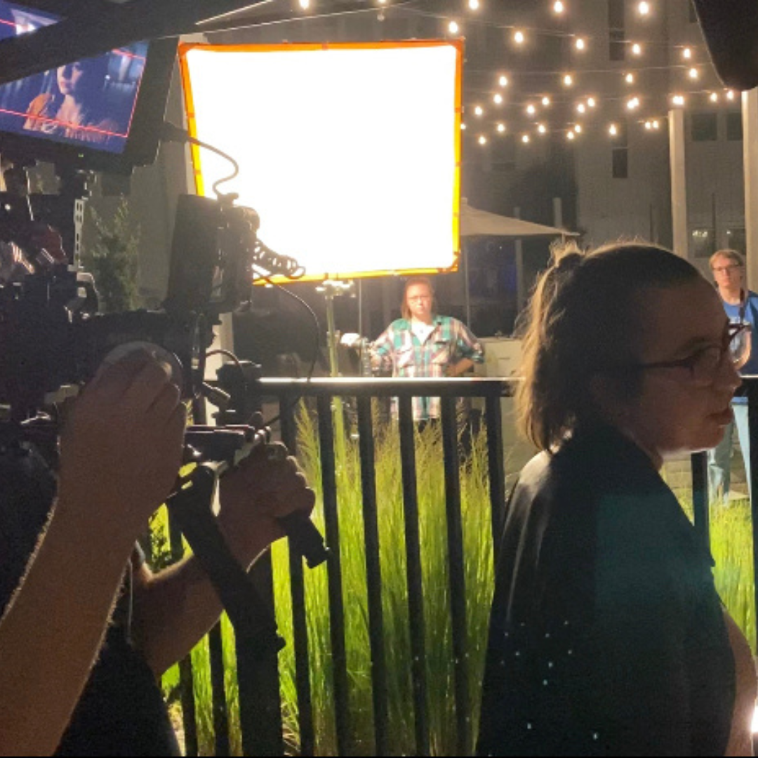 Nighttime outdoor scene with a woman in a black jacket and glasses standing near a black fence, illuminated by string lights. Several people are in the background, including a woman with a plaid shirt and glasses, and a large bright screen and camera