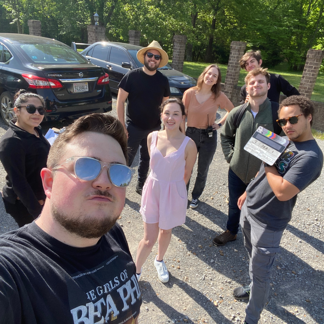 Group of nine young adults smiling outdoors on a sunny day, standing near parked cars, with green trees in the background.