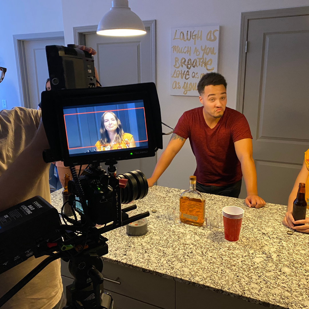 A man in a red t-shirt plays with a camera filming a woman who is smiling on a monitor. The scene is set in a kitchen with a granite countertop, a bottle of liquor, red plastic cups, and a person holding a beer. A wall sign reads "Laugh as much as yo