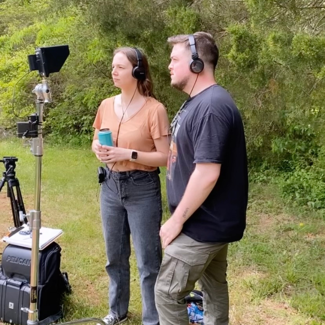 Two people standing outdoors on a film set, both wearing headphones and looking at a camera setup, with greenery in the background.