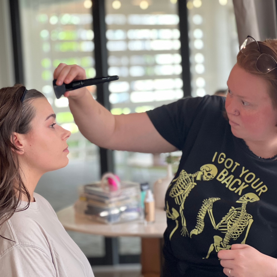 A makeup artist applying makeup on a woman with long brown hair, sitting indoors with natural light coming through slatted windows.