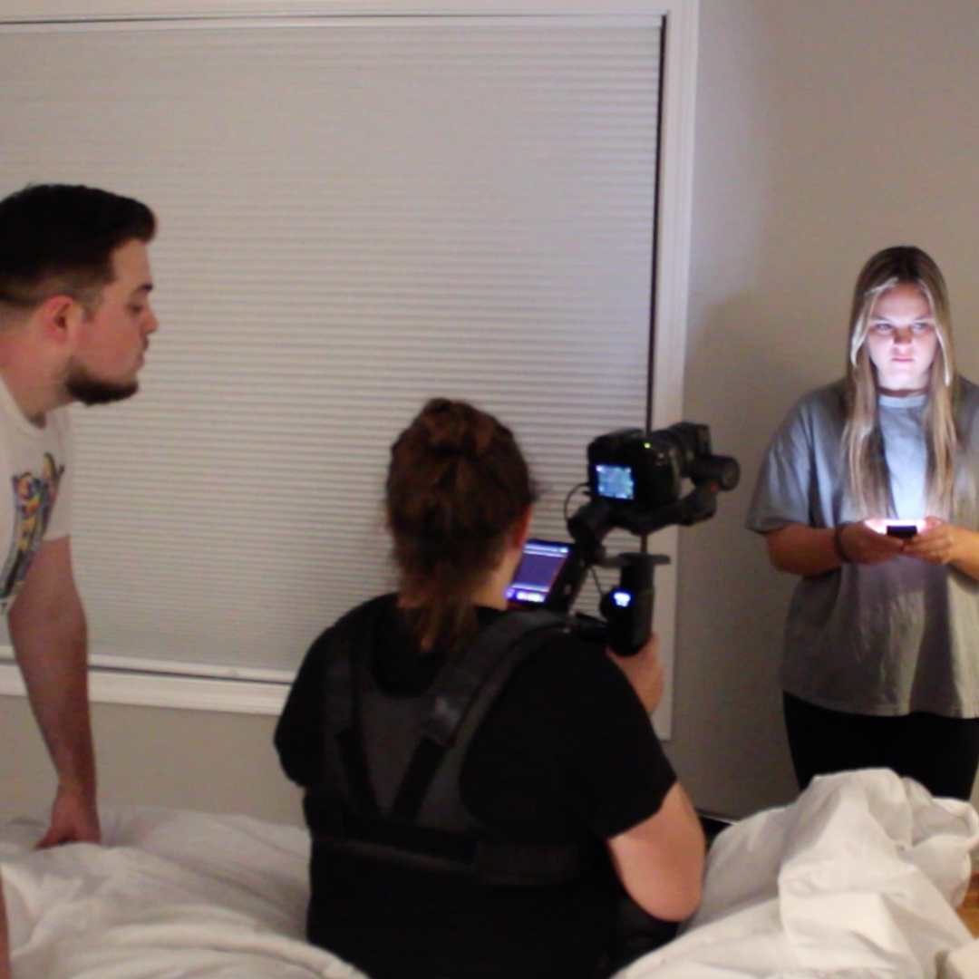 A young woman standing next to a bed, holding a phone with her face lit by the phone screen, as two people film her in a room.