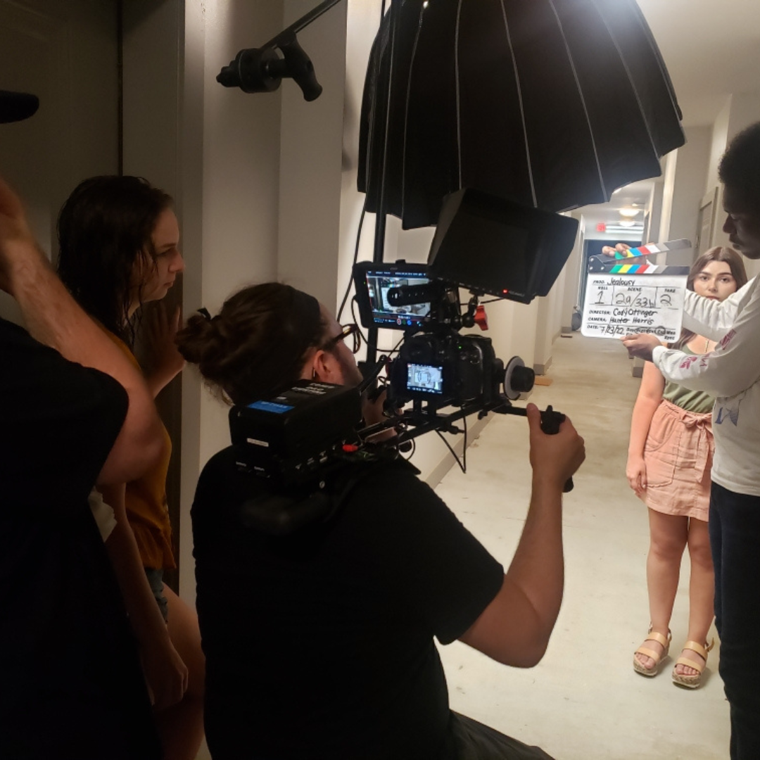 Filming scene with a camera operator, three women, and a clapperboard in a hallway.