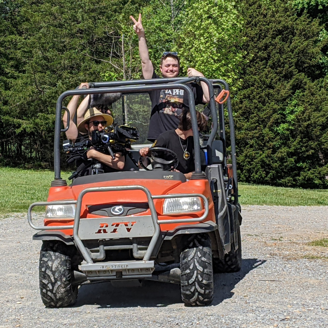 Three people riding on an orange utility vehicle on a gravel area in front of green trees. One person is standing with one arm raised and making a peace sign, while the others are seated, one wearing sunglasses and a wide-brim hat.