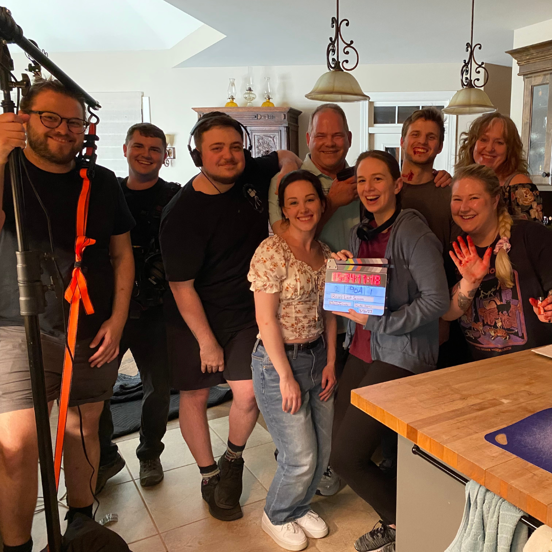 Group of nine people smiling and celebrating in a kitchen, some wearing headphones and holding a clapperboard, indicating a film or video production setting.