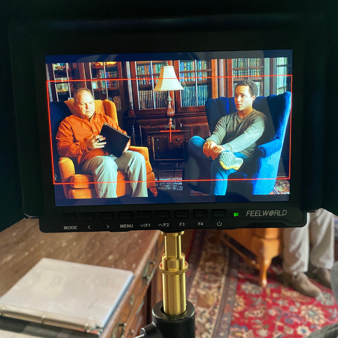 Film set monitor showing two men sitting in a library setting, one reading a tablet and the other sitting with crossed legs, engaged in conversation.