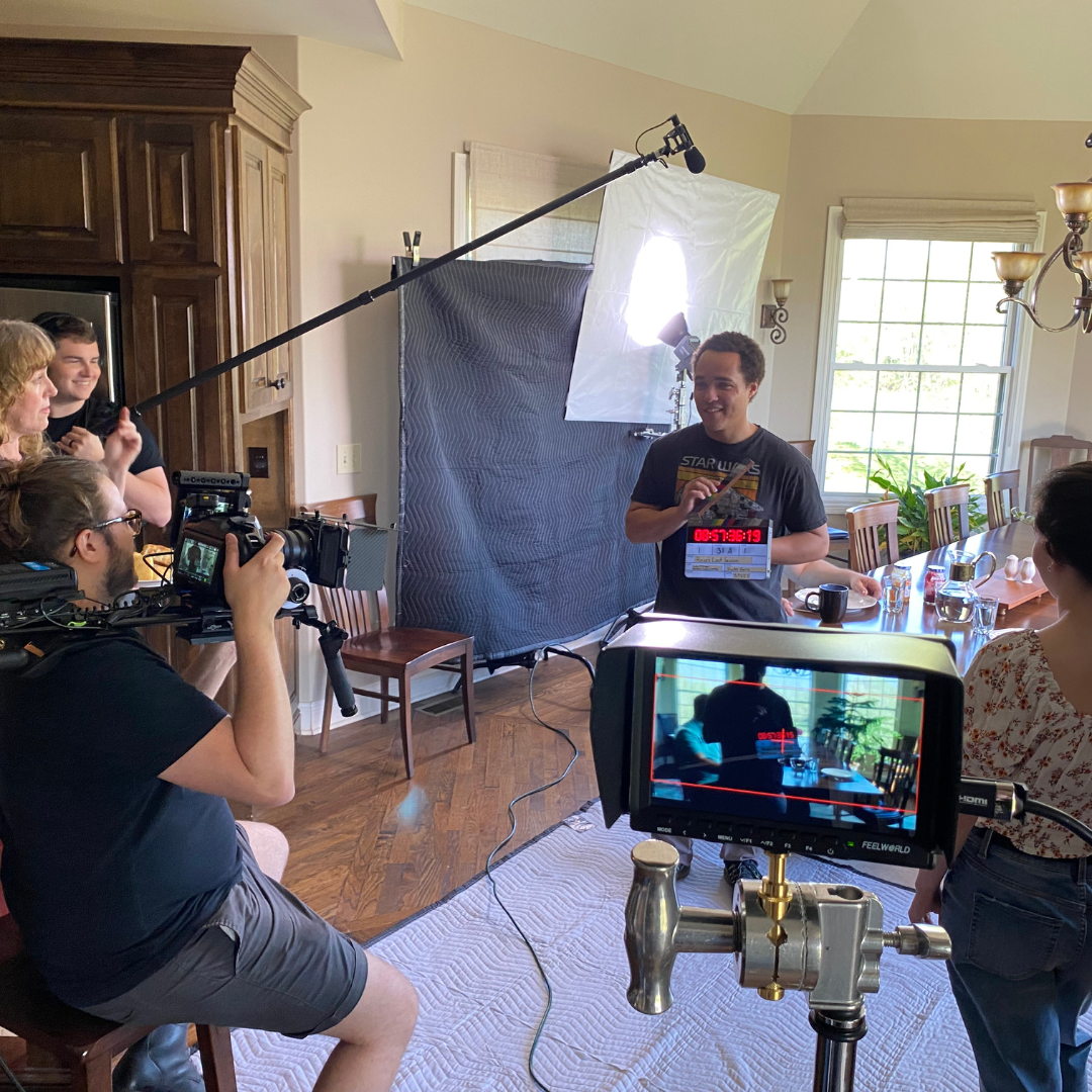 Filming scene in a dining room with a crew and actor, using lighting and camera equipment for a video shoot.