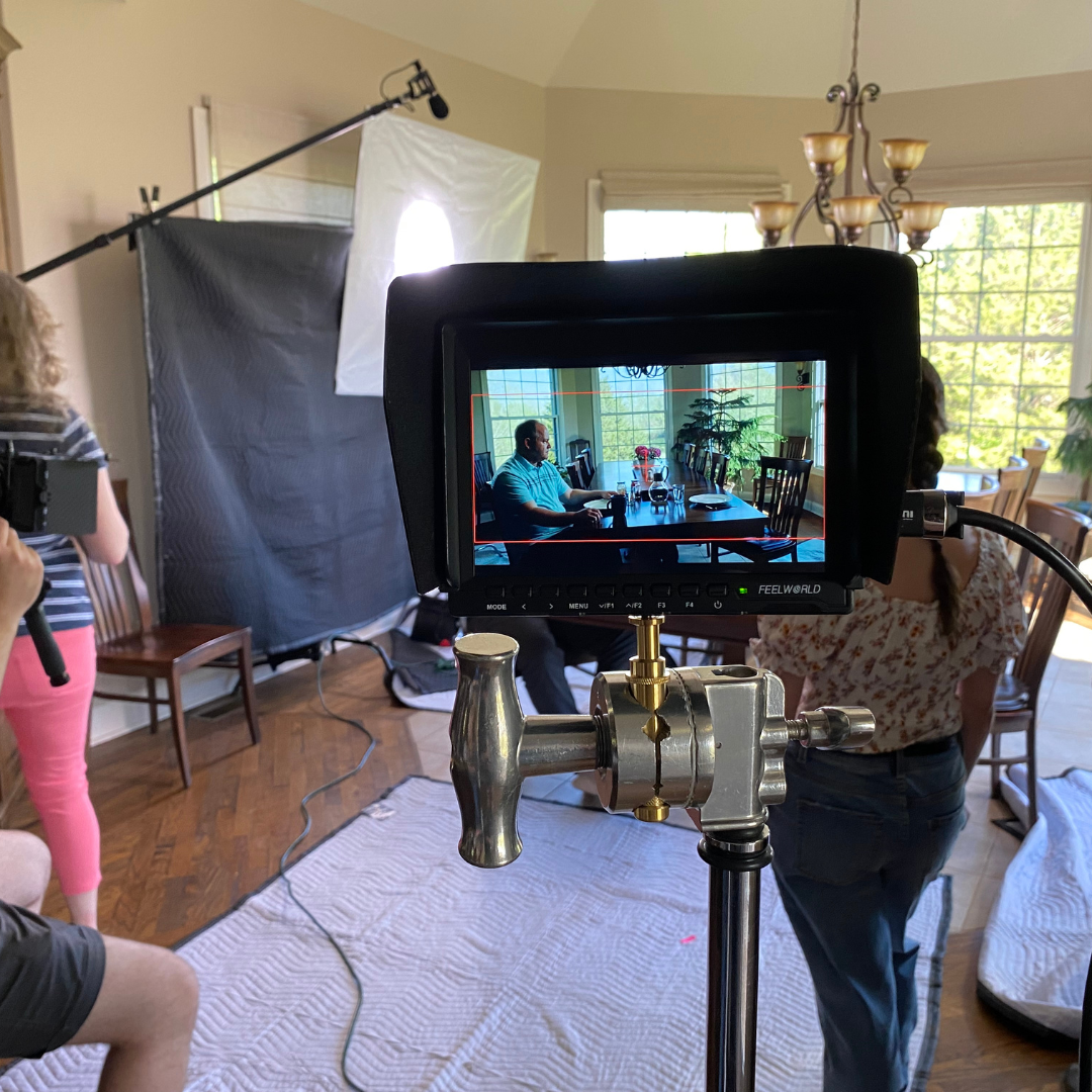 A filming setup capturing a man seated at a dining table in a bright room with large windows, surrounded by filming equipment and crew members.