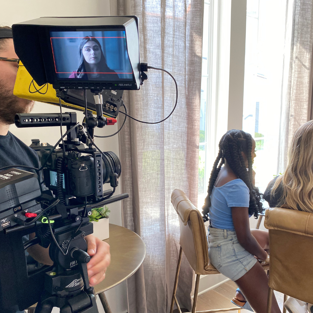 A woman sitting at a table, being filmed by a camera operator with a professional video camera. The scene is indoors, near large windows with curtains, during the daytime.
