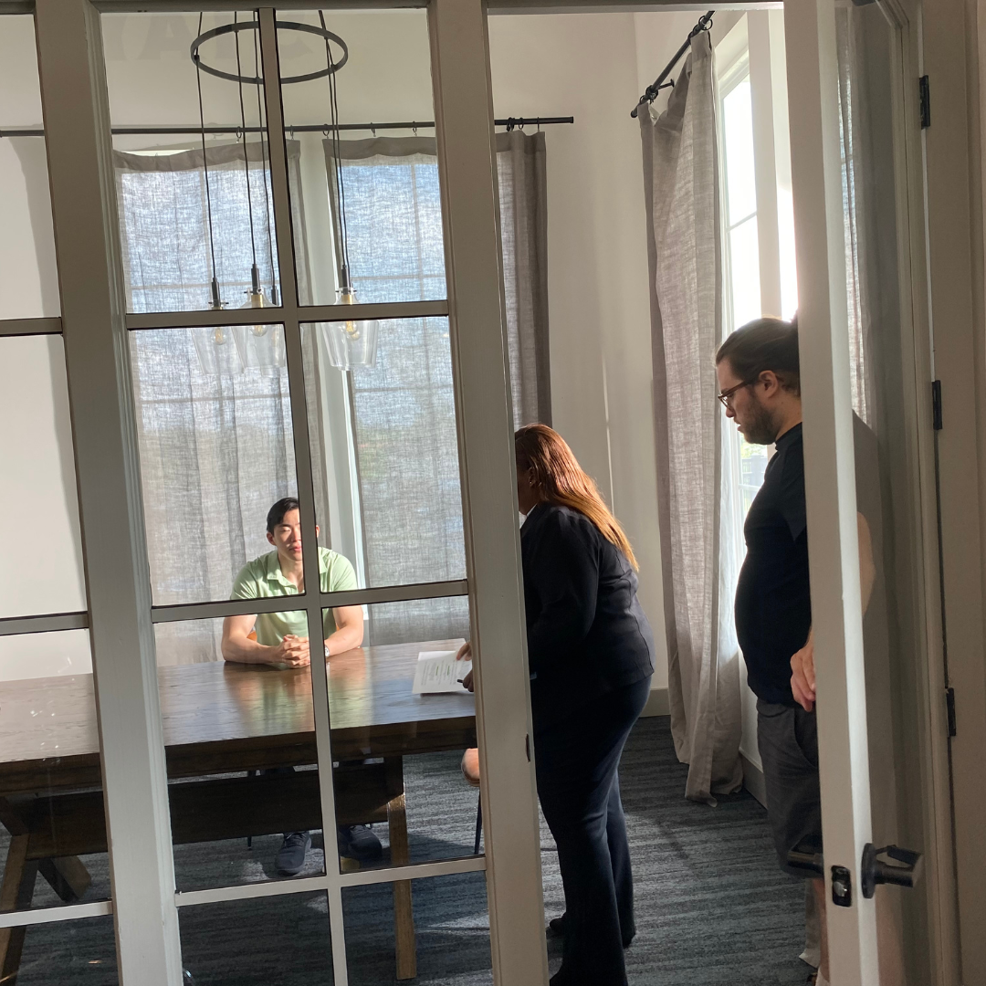 A person is sitting at a wooden table across from two standing individuals in a well-lit room with large windows, curtains, and a modern light fixture.