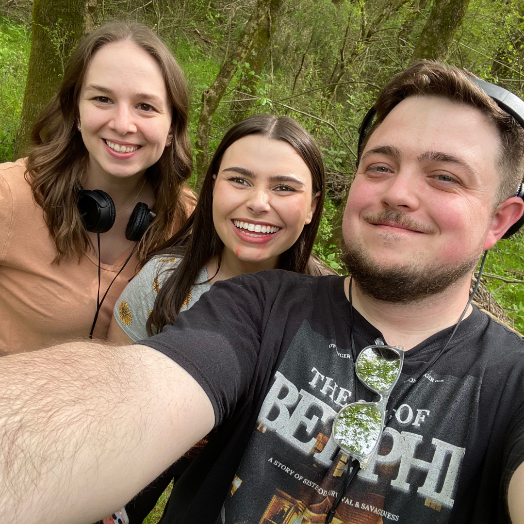 Three young adults smiling and taking a selfie outdoors in a wooded park, two women and one man, with headphones and sunglasses.