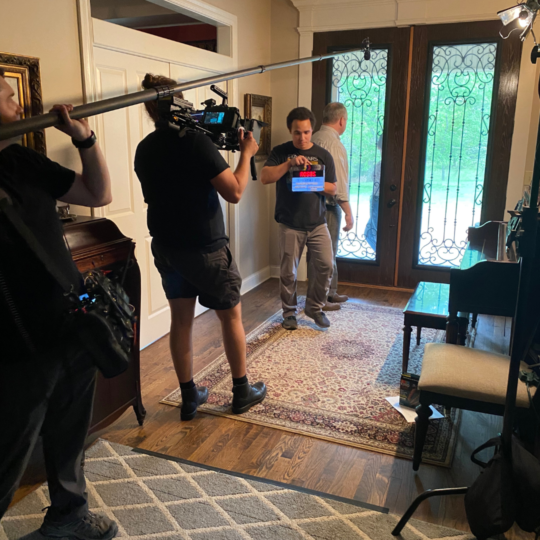 Behind-the-scenes of a film or TV production in a home with crew members, camera operator, and actor preparing for a shot near the front door with decorative ironwork on glass doors.