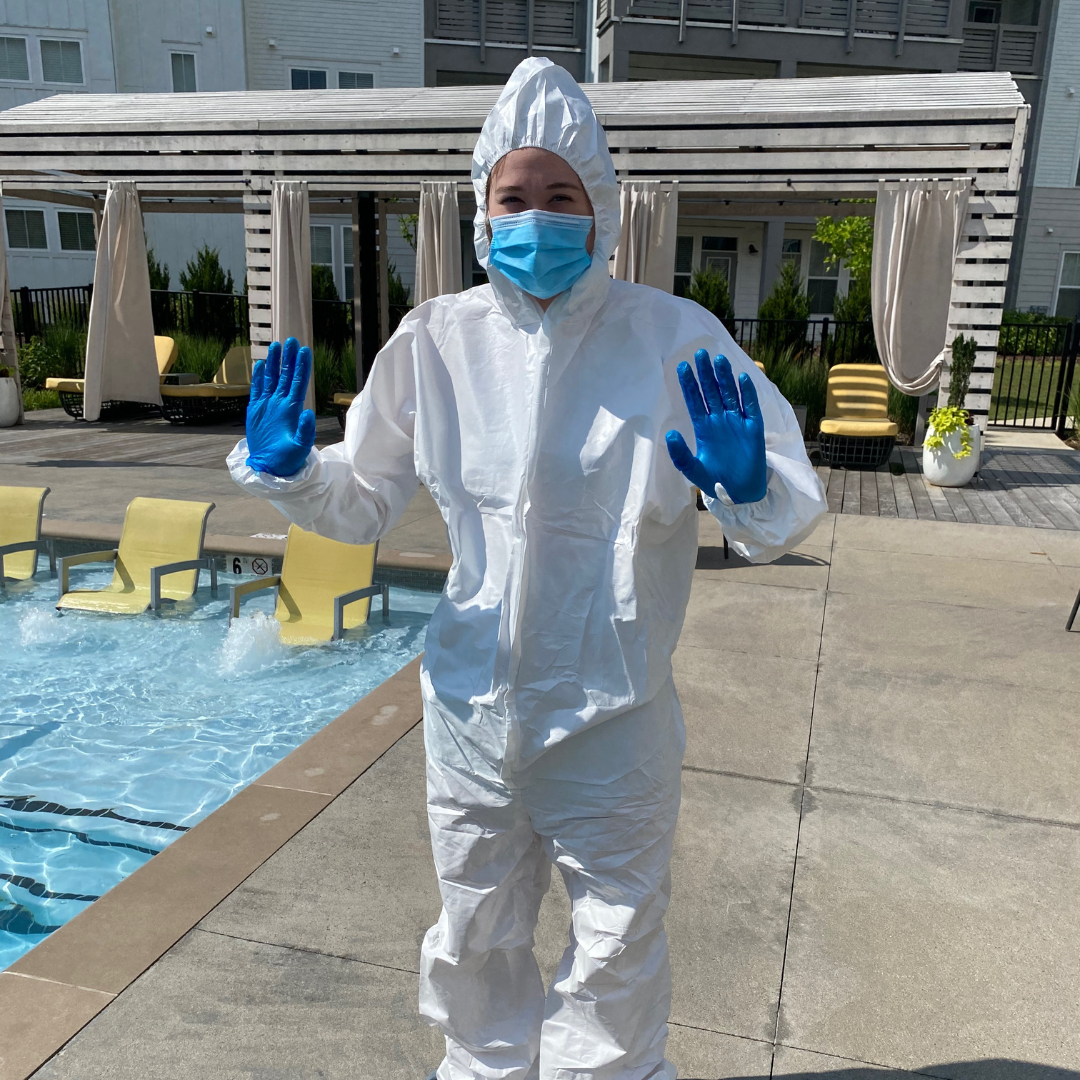 Person in protective gear, including a white hazmat suit, blue gloves, face mask, and surgical cap, standing by a swimming pool with pool chairs and a wooden cabana in the background.