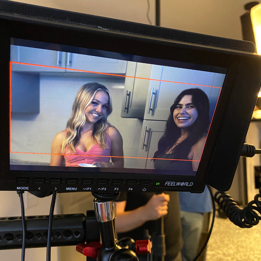 Two women smiling and talking, seen through a monitor on a film or video set, with kitchen cabinets in the background.