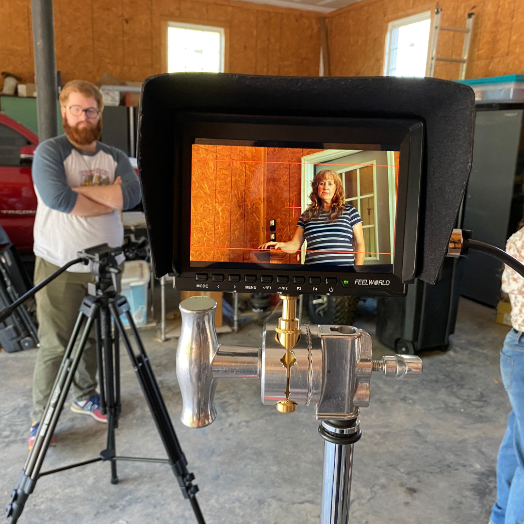 A video camera's viewfinder showing a woman in a striped shirt standing in front of a woman with long hair. The background includes a doorway and wooden walls.