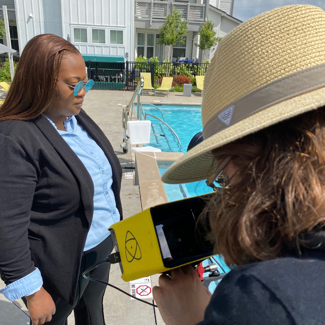 A woman in a black blazer and blue shirt stands poolside looking down as another woman in a wide-brimmed straw hat and glasses uses a handheld device, possibly for testing or measurement, near a swimming pool with an apartment or condo complex in the