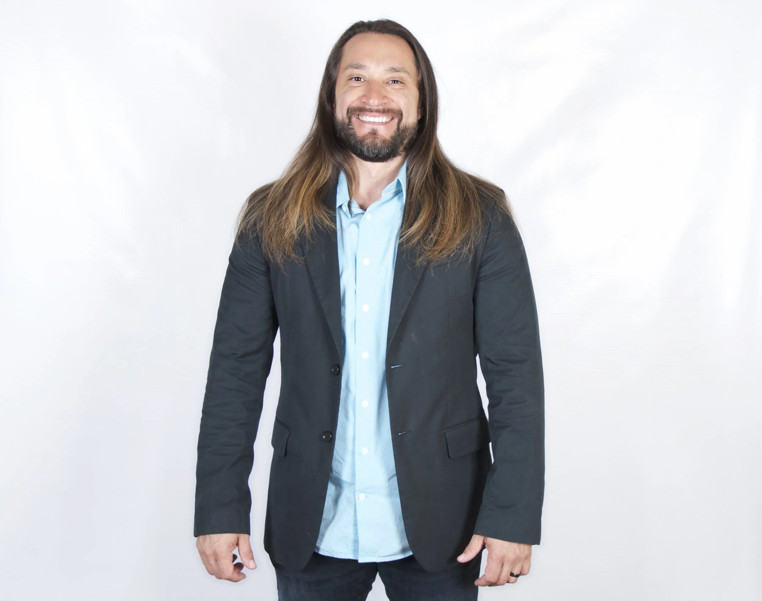 Smiling man with long brown hair and beard wearing a dark blazer, light blue shirt, and black pants, standing against a plain white background.