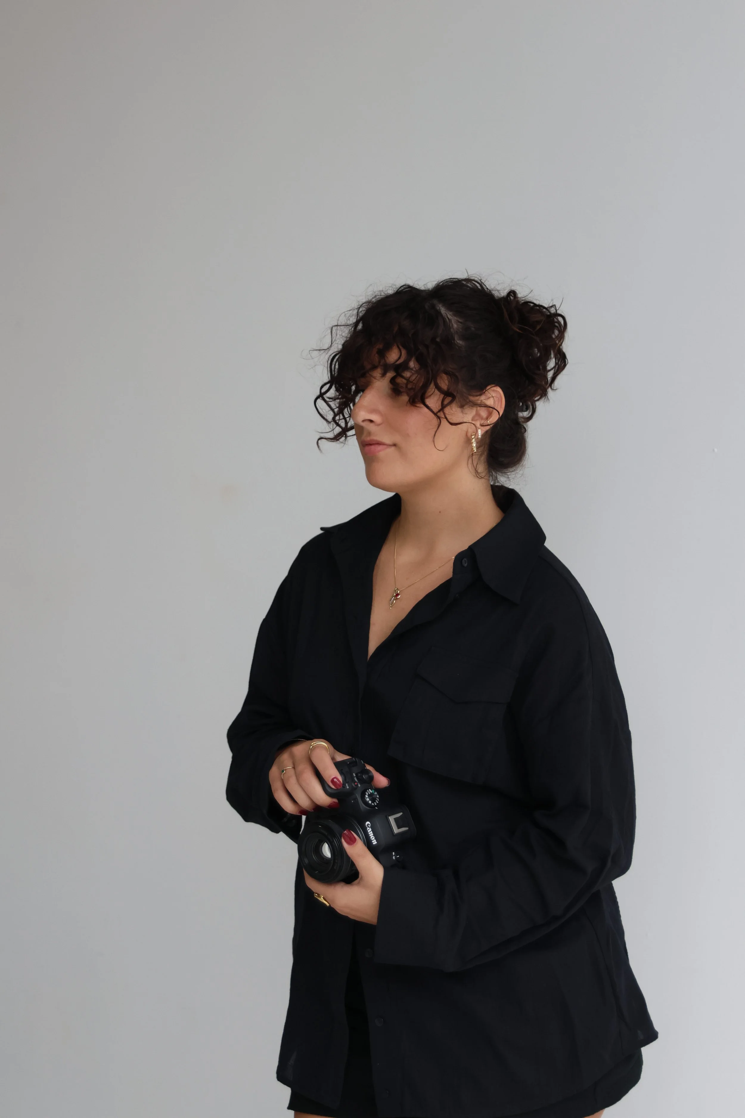 A woman with curly hair holding a camera, wearing a black shirt and jewelry, standing against a plain white wall.