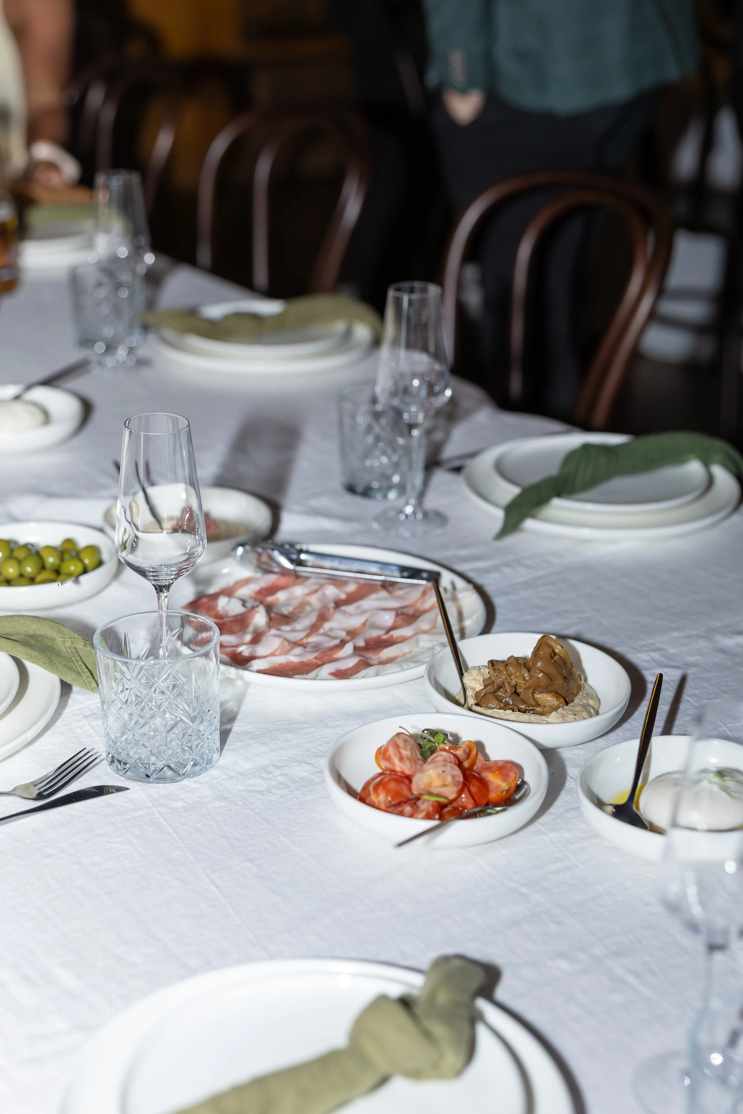 Table set for a meal with plates, glasses, and bowls containing sliced tomatoes, cold cuts, and a casserole dish, with green napkins on plates.
