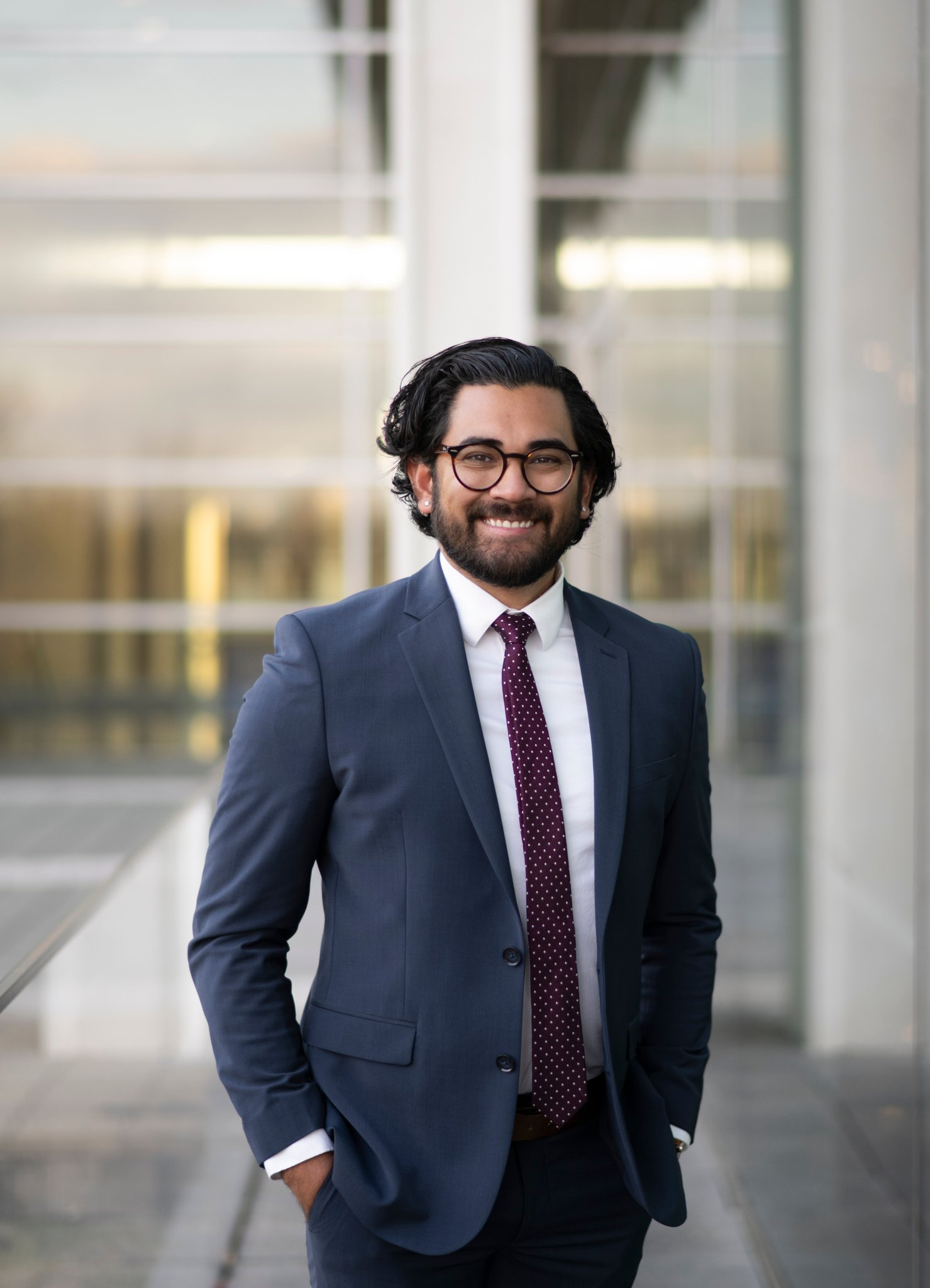 Professional man wearing a navy suit and tie, smiling while standing in front of a modern glass building.