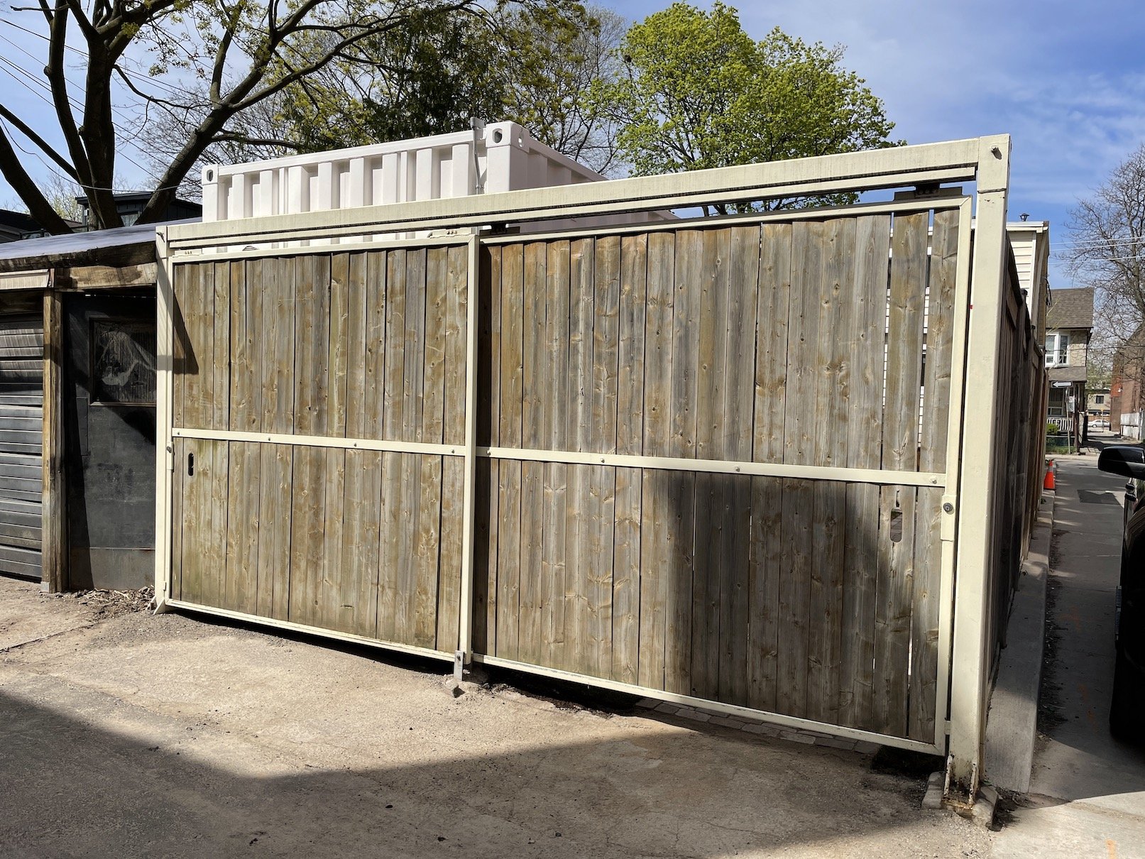 A wooden sliding gate on a concrete driveway, with a white metal container on top. A gray building with a garage door is on the left, and other houses and trees are visible in the background.