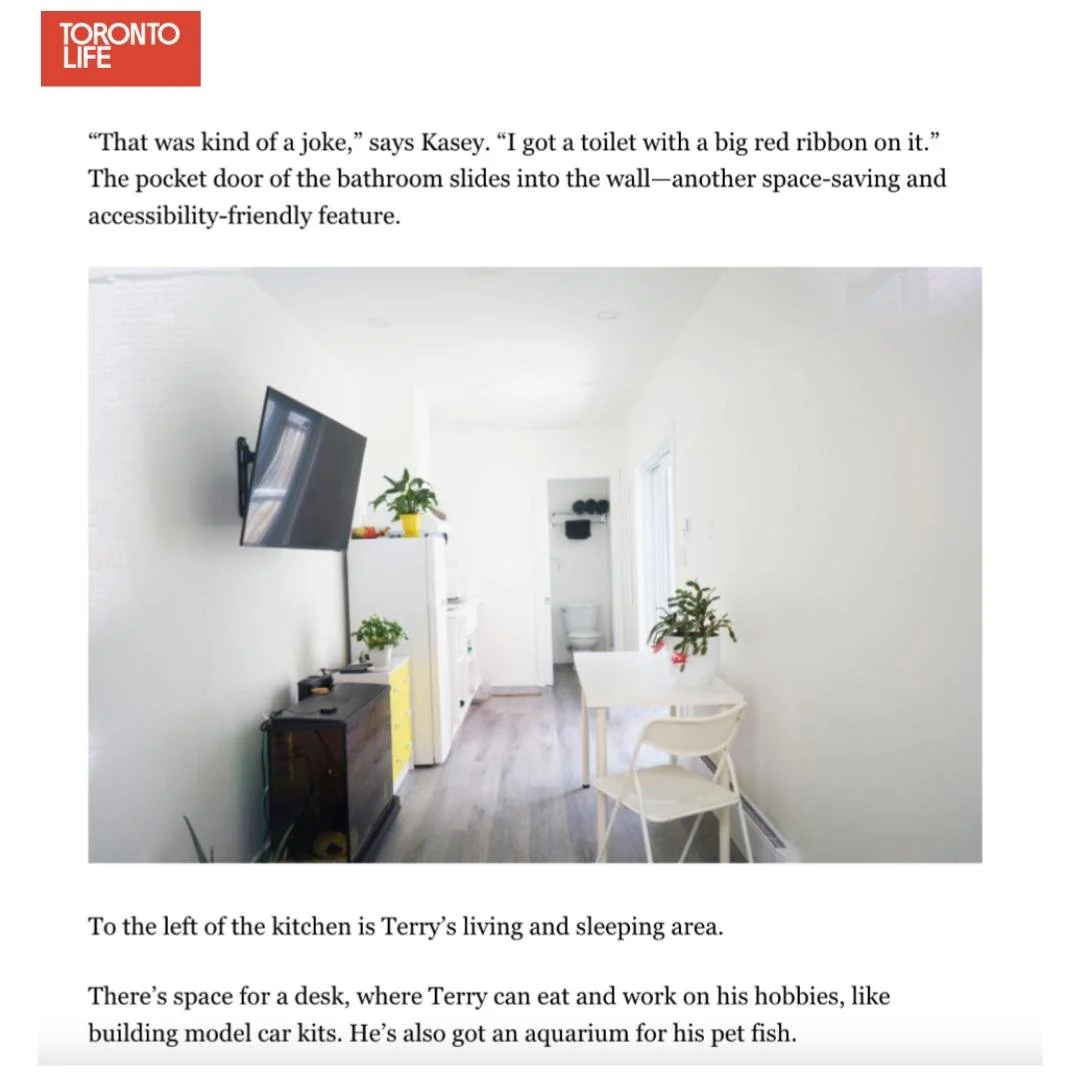 A small, minimalist living and sleeping area with a wall-mounted television, a white fridge, a small yellow cabinet, and a white table with chairs. There are plants and a view of a bathroom with a toilet in the background.