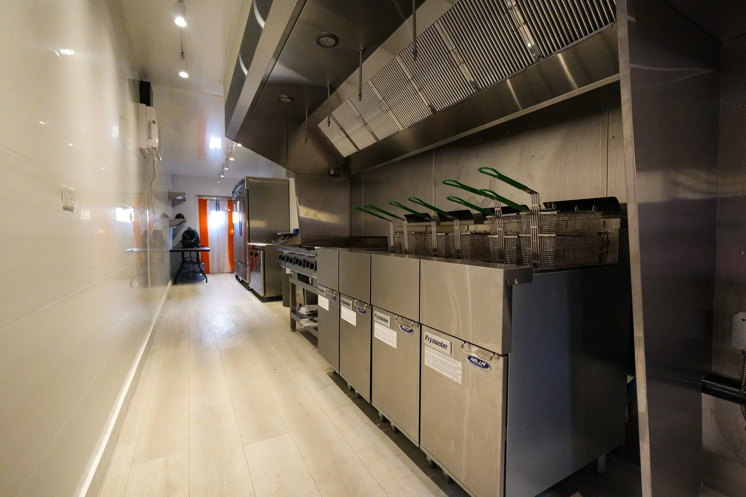Empty commercial kitchen with stainless steel equipment, fryers with green-handled baskets, stove, oven, and table in the background.