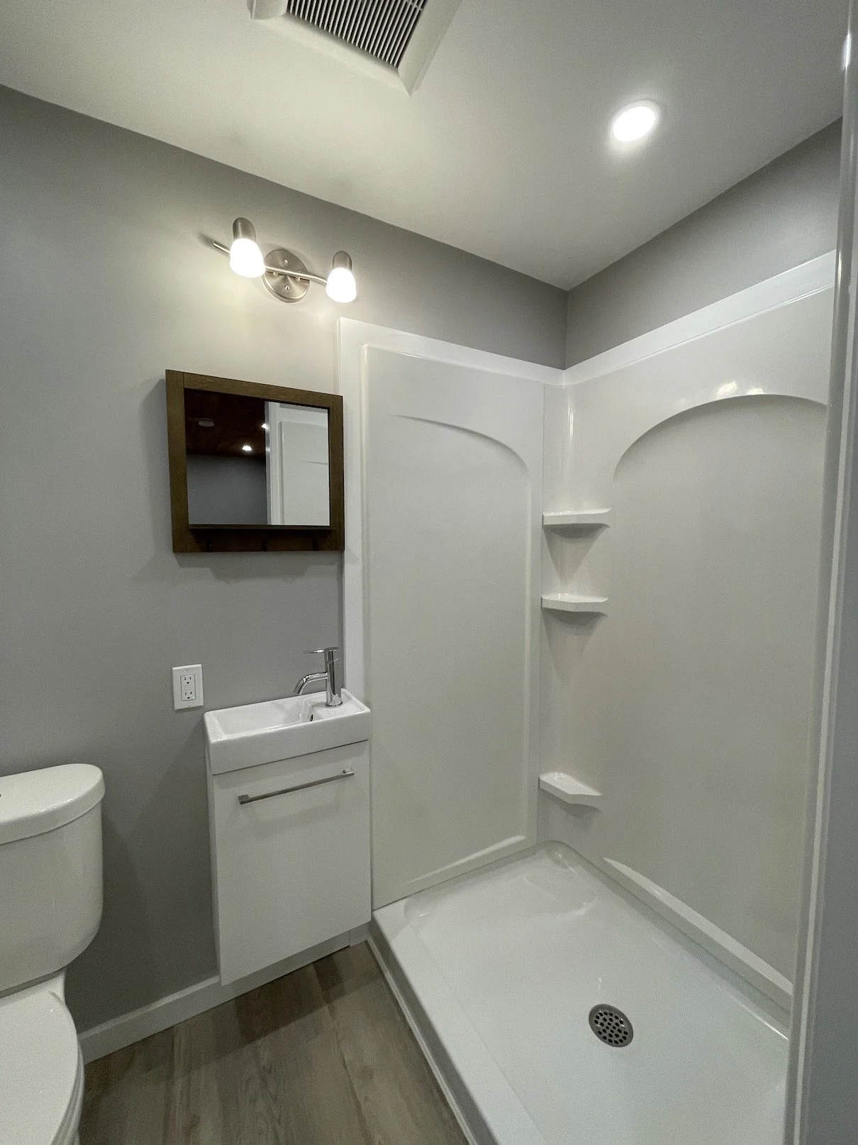 Modern bathroom featuring a shower with built-in shelves, small white sink with a cabinet, a mirror, a light fixture, a toilet, and wood-look flooring.