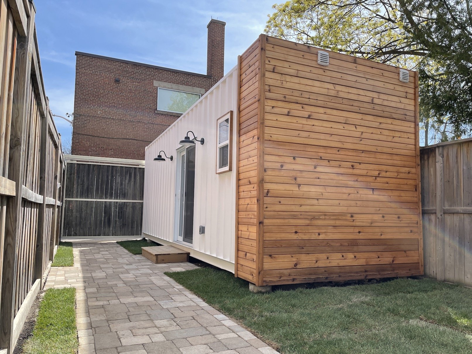 Small backyard with a paved walkway, wooden privacy fence, and a tiny white building with black outdoor lights and a window, adjacent to a larger brick building.