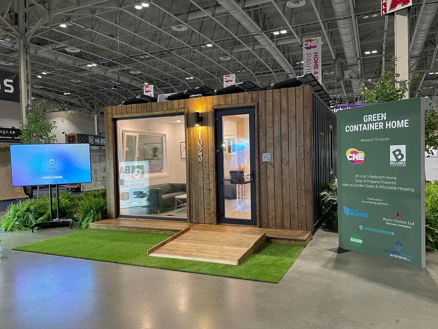 A small, modern model home made from repurposed shipping containers with wooden siding and large windows, displayed indoors at a home and garden show.