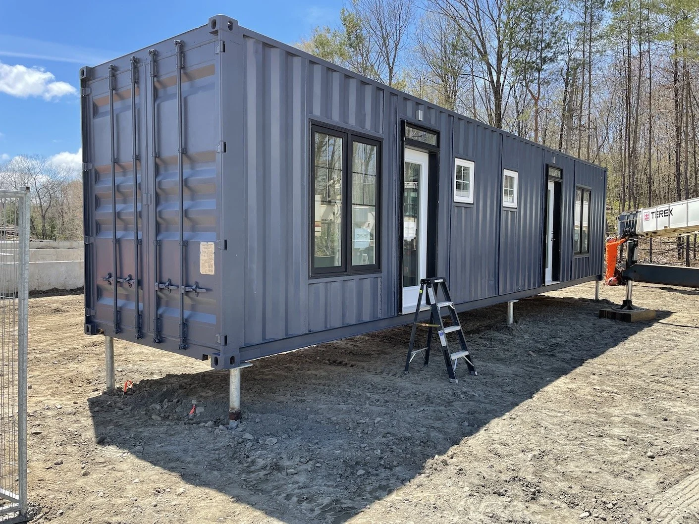 A shipping container converted into a tiny home, featuring multiple windows and a door, set on a construction site with trees in the background.
