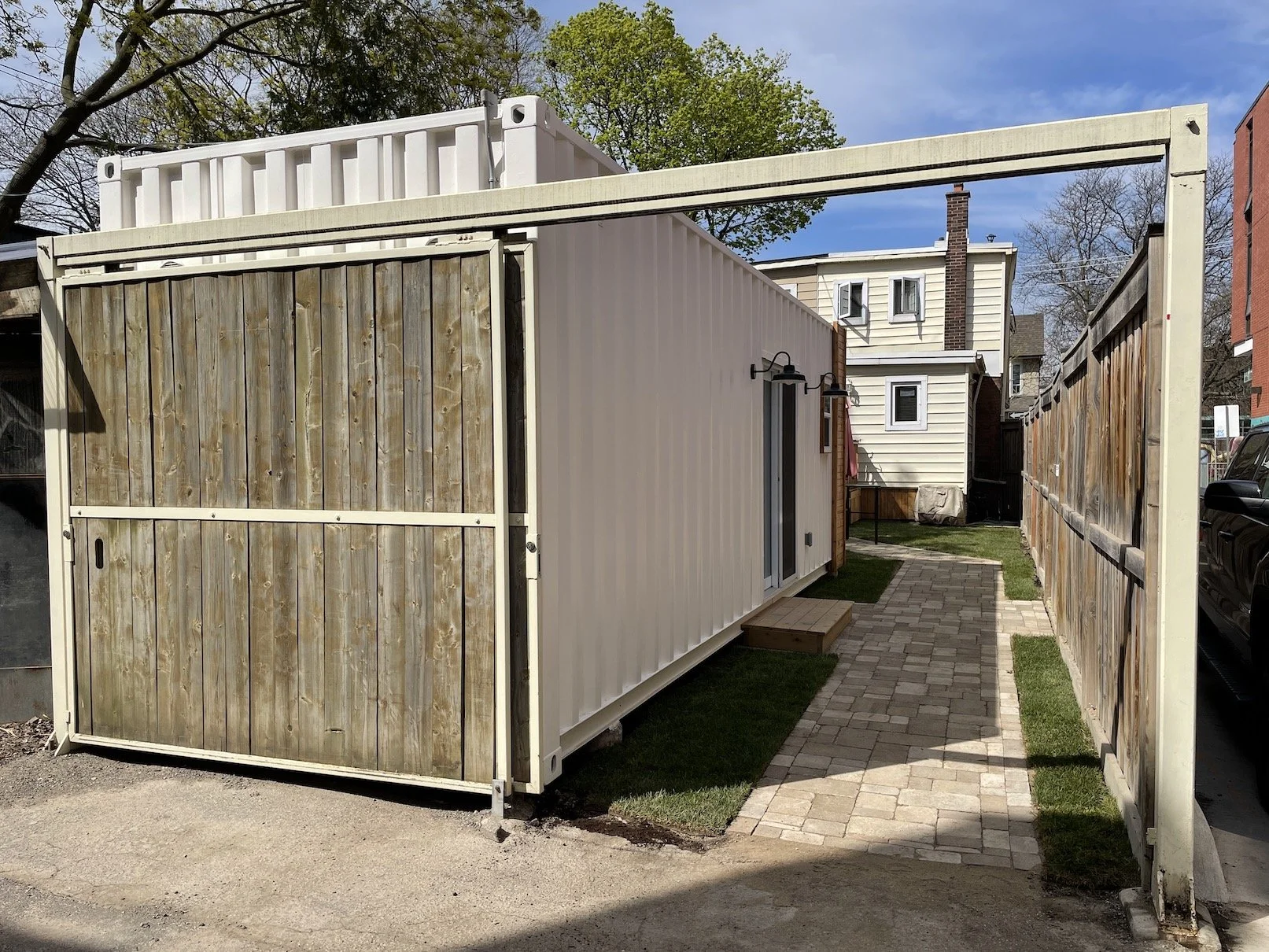 A small backyard with a white shipping container house, a wooden gate, and a stone pathway. There are small grassy areas and neighboring houses visible in the background.