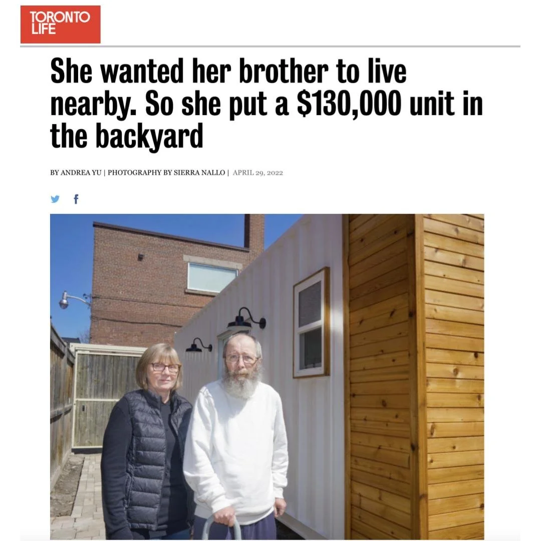 A woman and an elderly man standing outside next to a tiny backyard house with a mix of white and wooden exterior walls, under a clear blue sky.