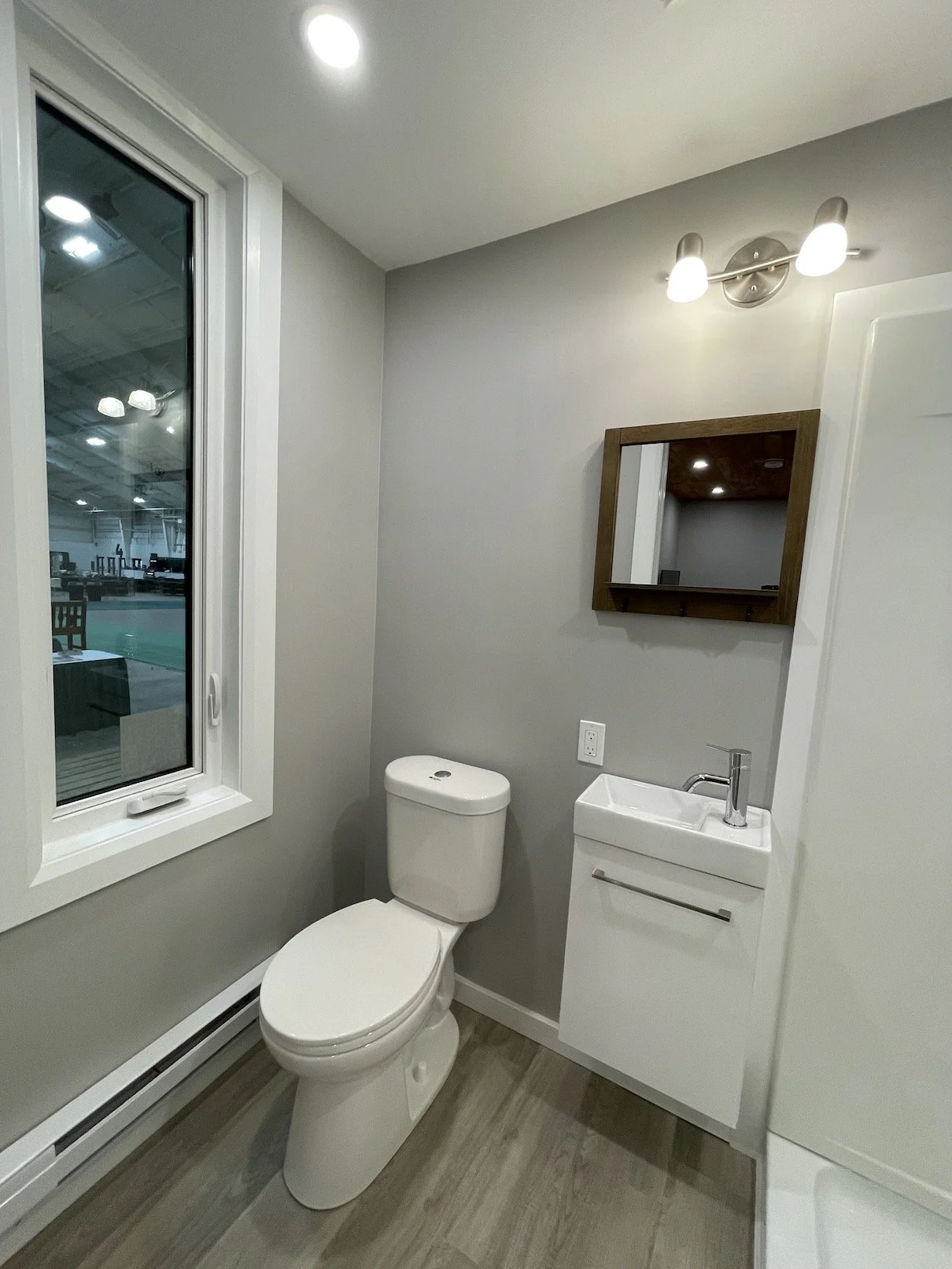Small modern bathroom with a white toilet, small sink with a cabinet, a mirror, a small wall-mounted light fixture, and a window showing an indoor gym or sports facility.