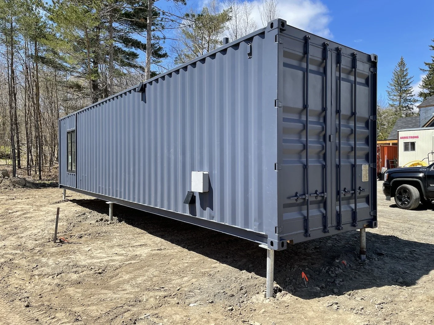 Gray shipping container with a window, set up on supports, outdoors on dirt ground with trees and a clear sky in the background.