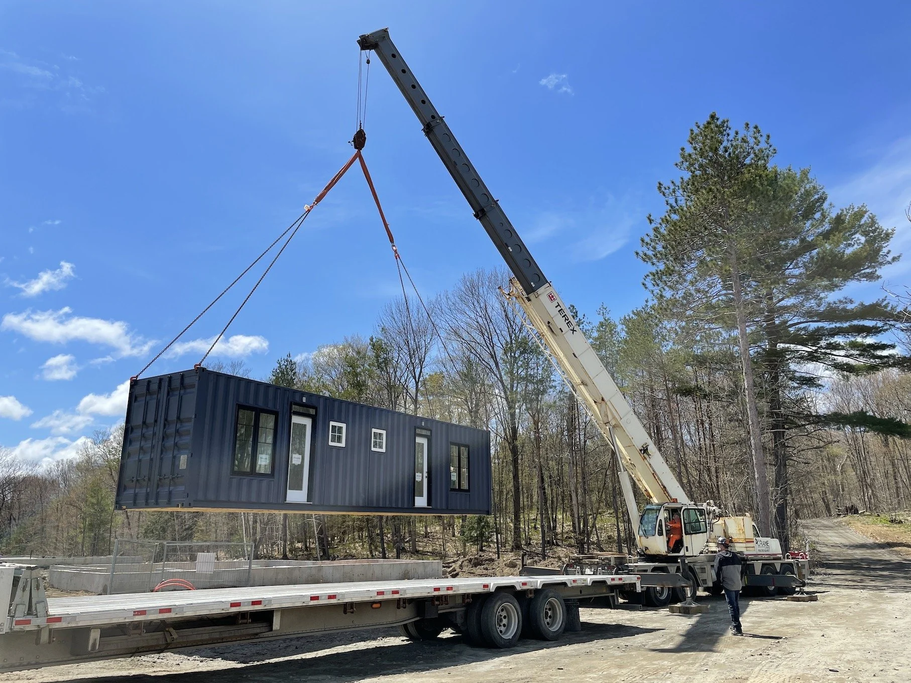 A large crane lifting a tiny prefabricated home onto a flatbed truck in a wooded outdoor area.