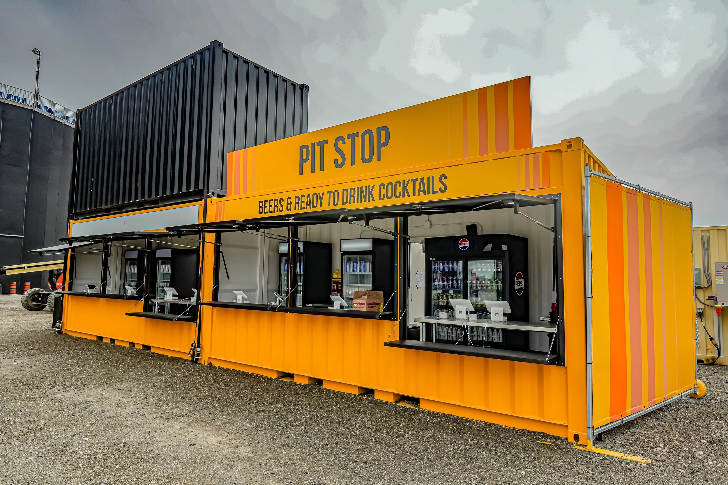 A bright yellow mobile food stand labeled 'PIT STOP' that offers beers and cocktails, with multiple service windows and refrigerated beverage coolers inside. The stand is situated on a gravel lot with some industrial buildings in the background.