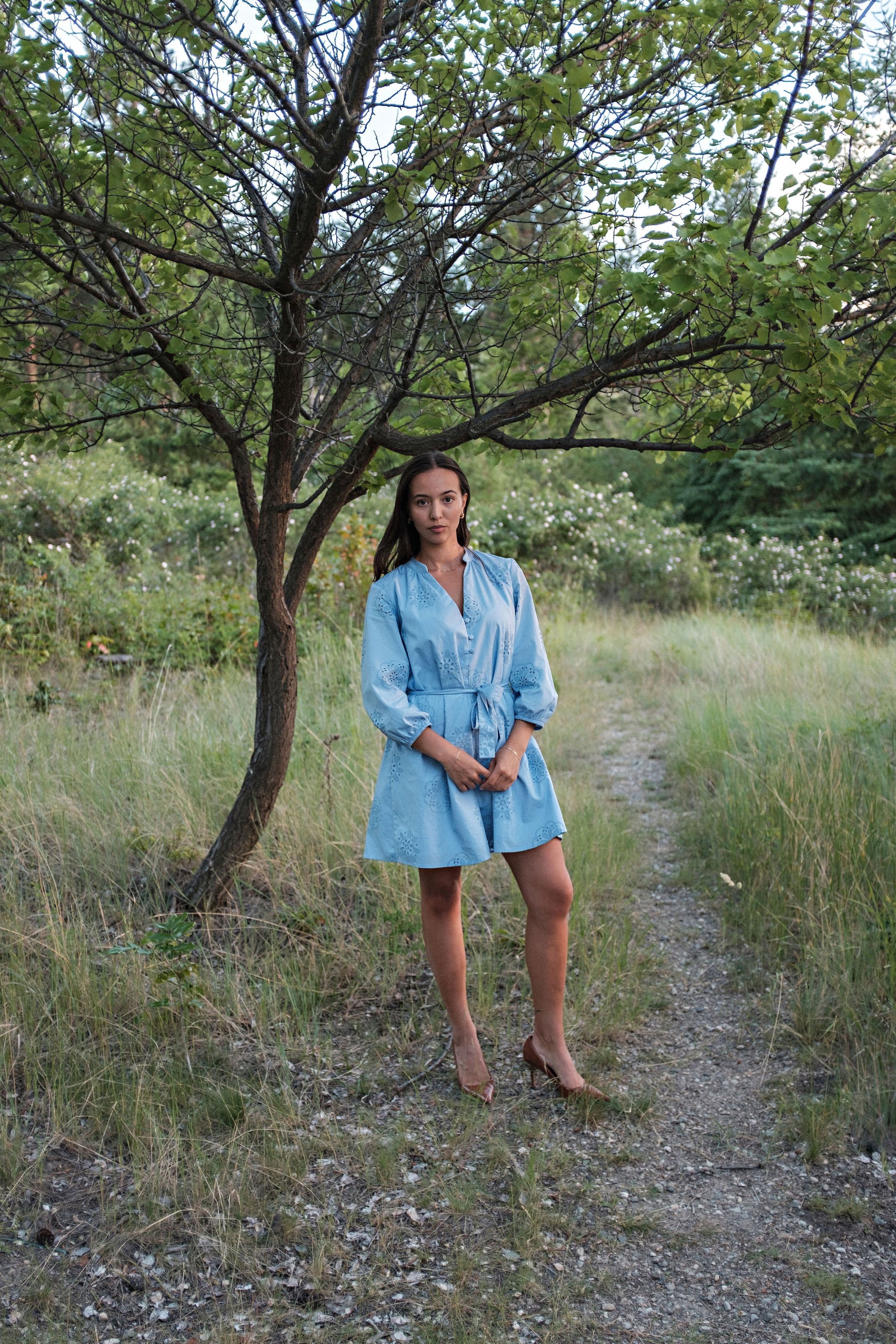 A woman in a light blue dress and high heels standing on a dirt path next to a tree in a grassy outdoor setting.