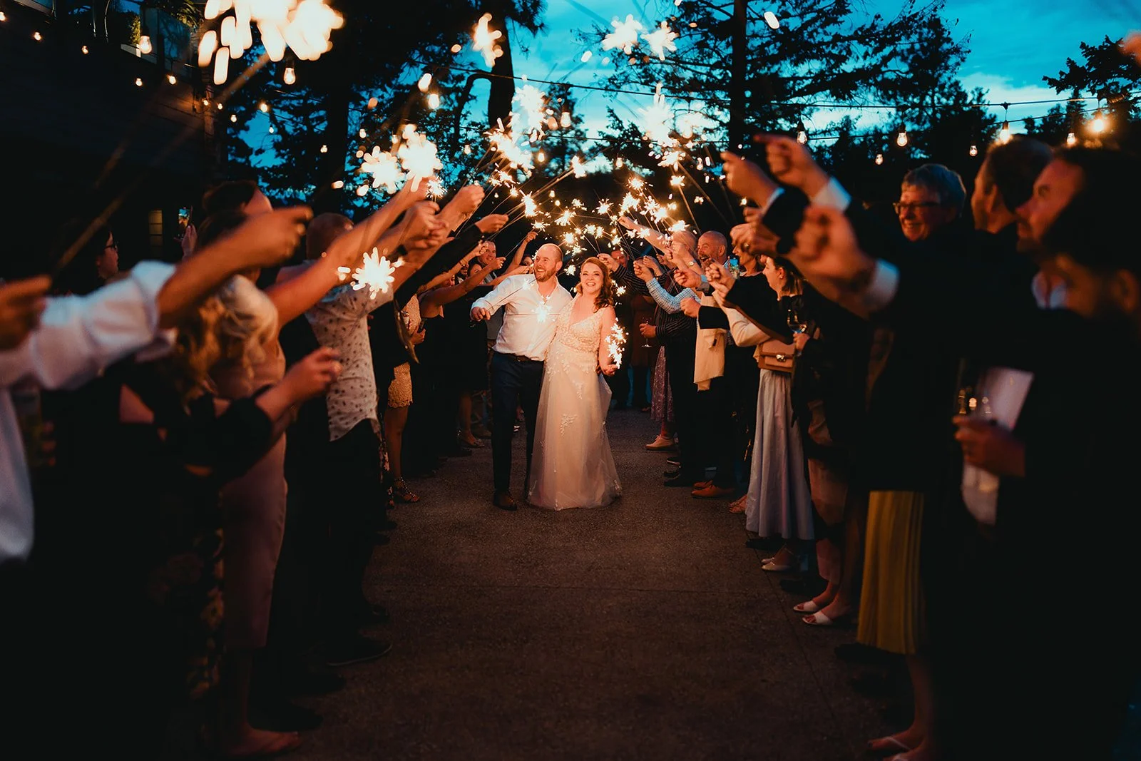 Wedding celebration with a bride and groom walking through a sparkler tunnel at dusk, surrounded by friends and family.