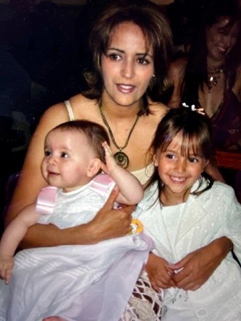 A woman holding a baby on her lap with a young girl beside her, all smiling and dressed in light-colored clothing at a social gathering.