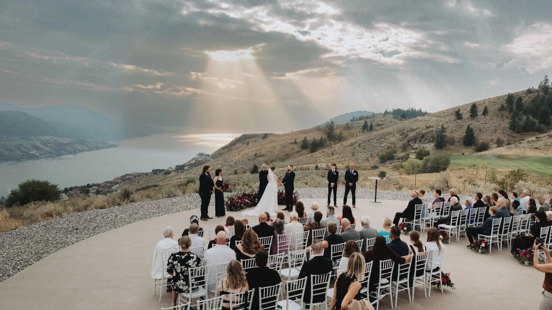 Outdoor wedding ceremony on a hill overlooking a lake and mountainous landscape, with a bride and groom exchanging vows, surrounded by guests seated in white chairs.