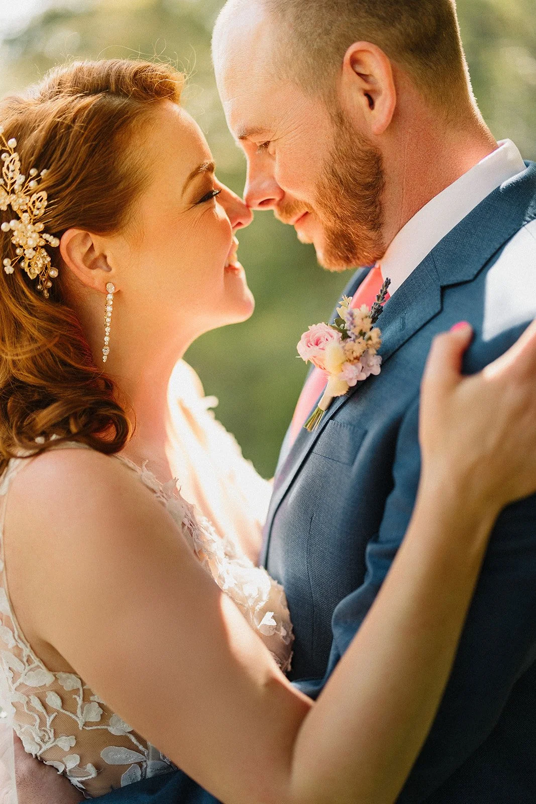 A bride and groom are close together, touching noses, smiling, and embracing each other outdoors on a sunny day.