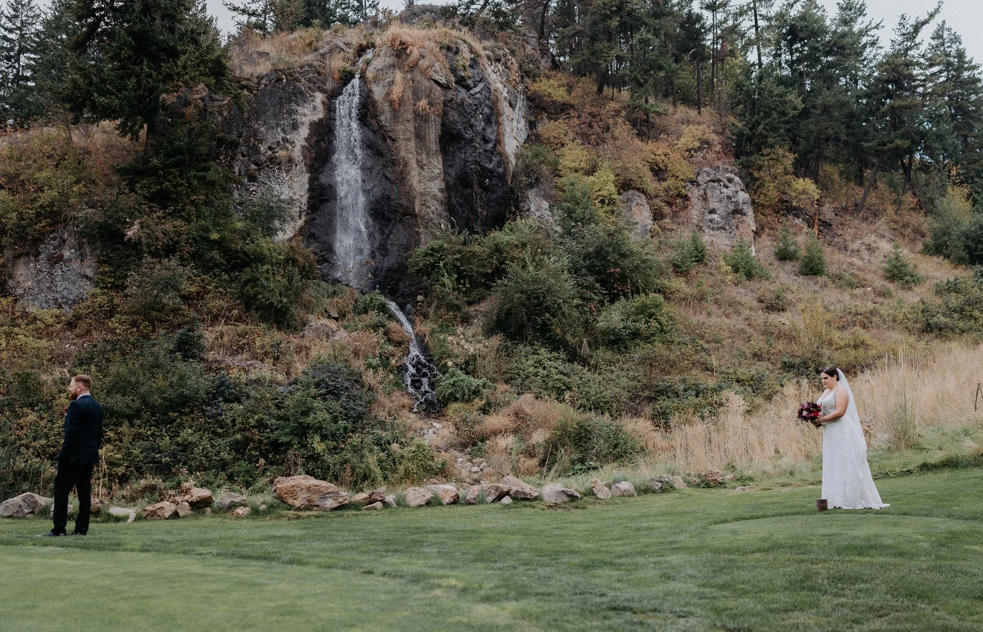 A bride in a white wedding dress holding a bouquet and a groom in a dark suit standing apart on a grassy area near a waterfall and wooded hillside.