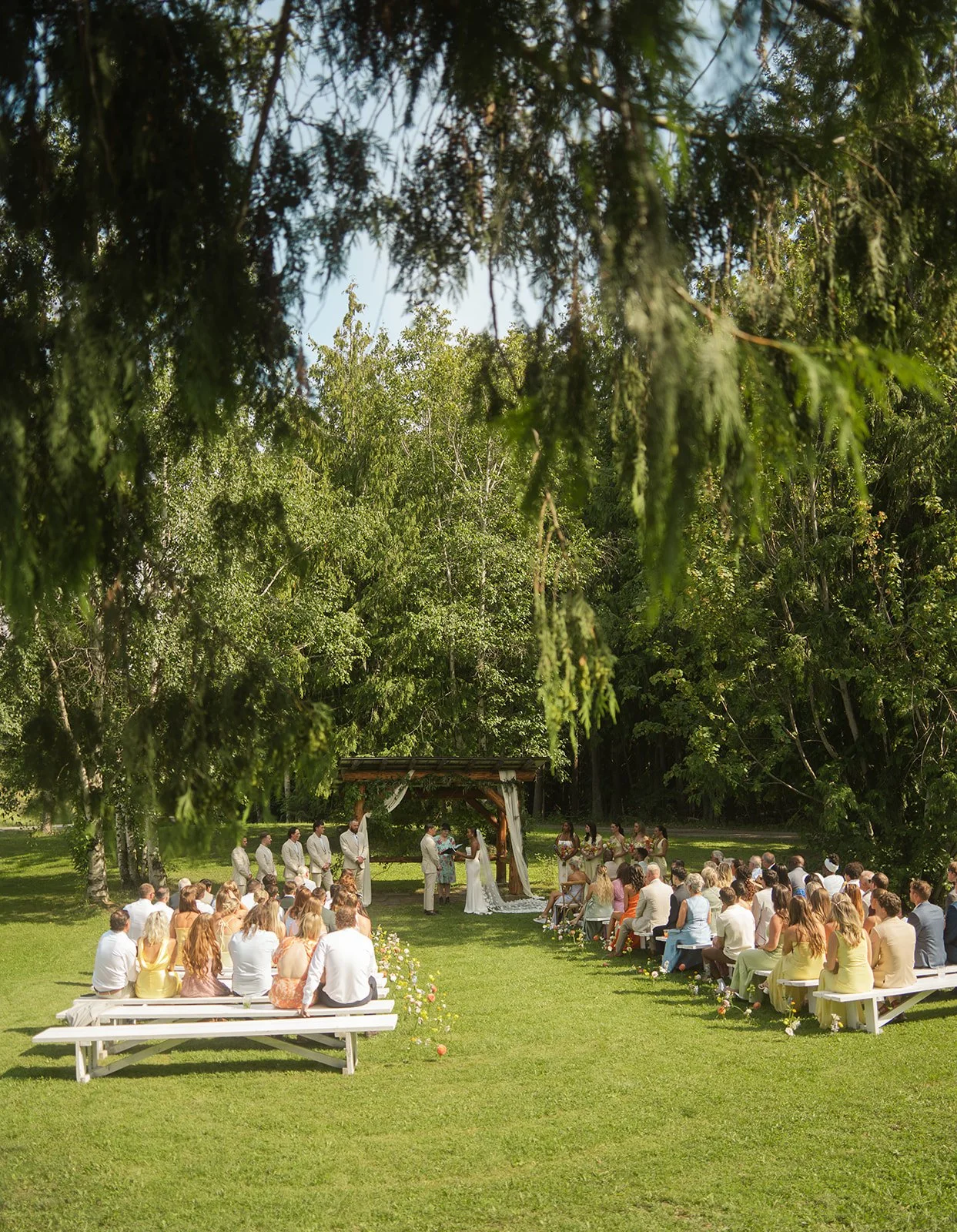 Outdoor wedding ceremony with guests seated on benches, officiants and bride and groom at altar surrounded by trees.