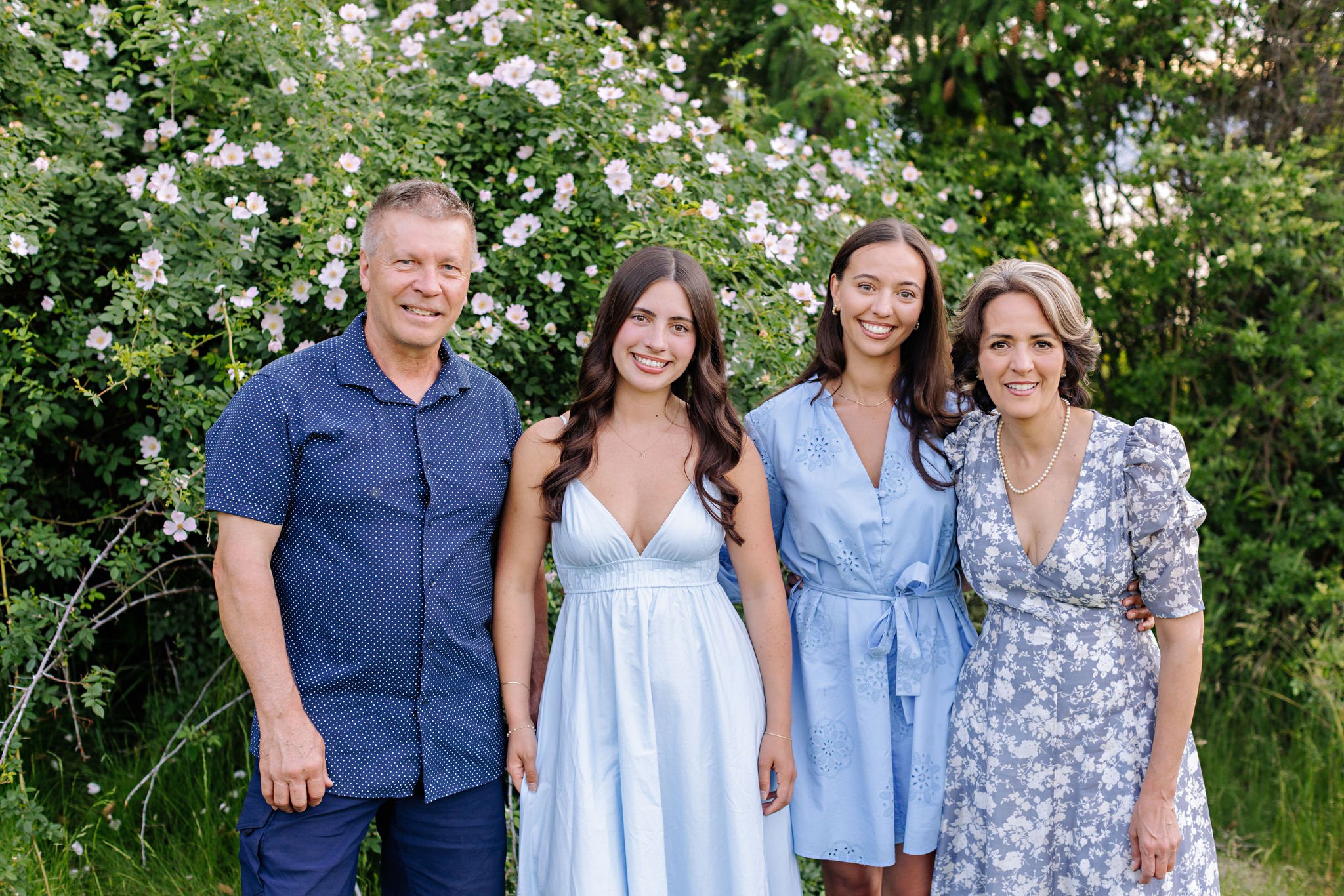 A family of four standing outdoors in front of blooming bushes. From left to right: a man, a young woman, a young woman, and an older woman. All are dressed in blue or white clothing and smiling.