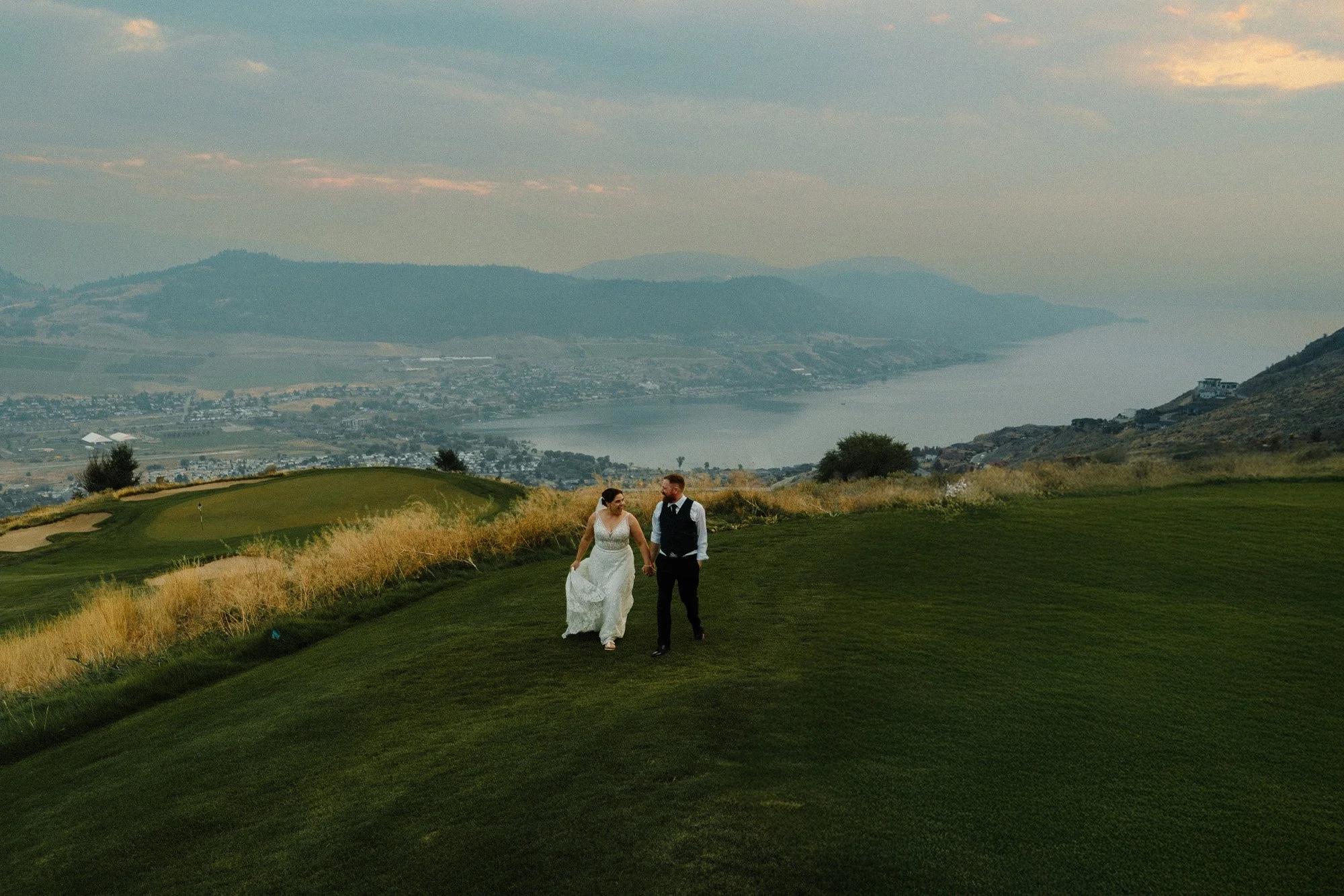 A newlywed couple walking hand in hand on a golf course with a scenic view of a lake and mountains in the background.