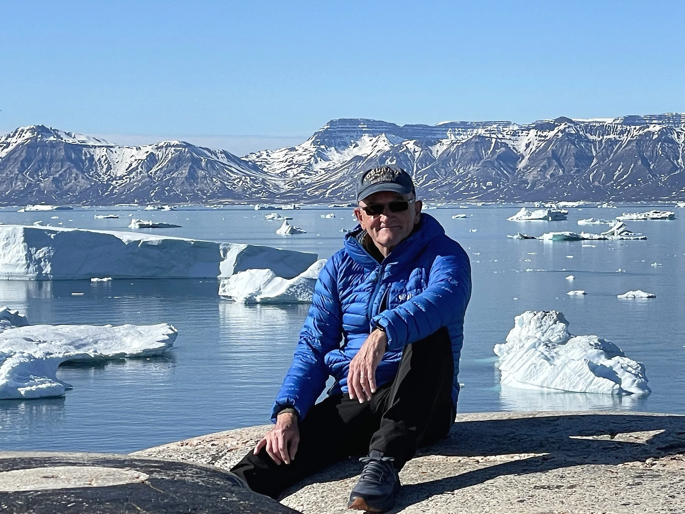 A man sitting on a rock near a body of water with icebergs, mountains, and clear sky in the background.